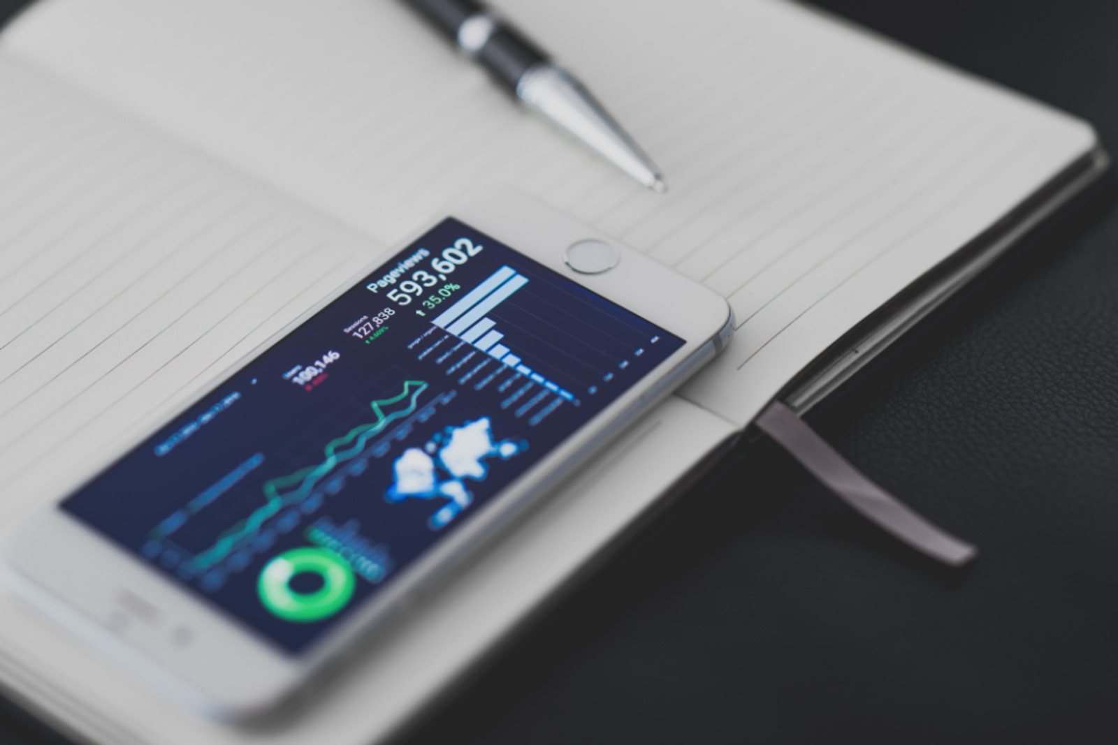 Смартфон с ярким экраном-дашбордом и графиками лежит на открытом блокноте рядом с ручкой